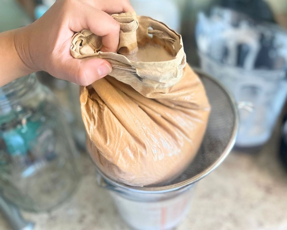 Full nut milk bag straining almond milk over a colander on top of a measuring cup