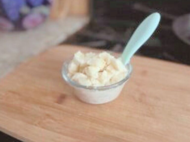 A glass bowl with cooked oatmeal with a small spoon dipped in it, placed on a wooden board on a stove.