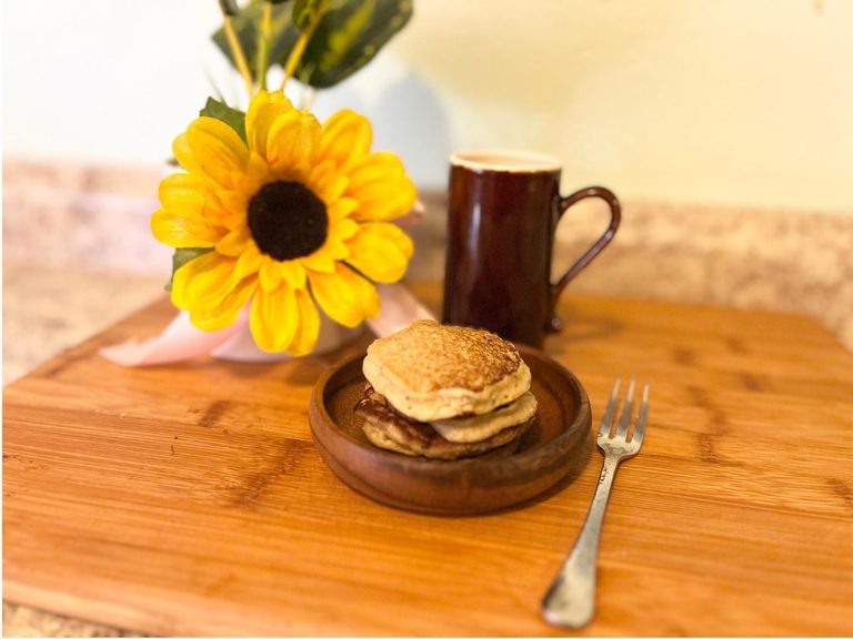 Yellow daisy flower, brown tea cup, pancake stack on a wooden plate, silver fork on top of cutting board.
