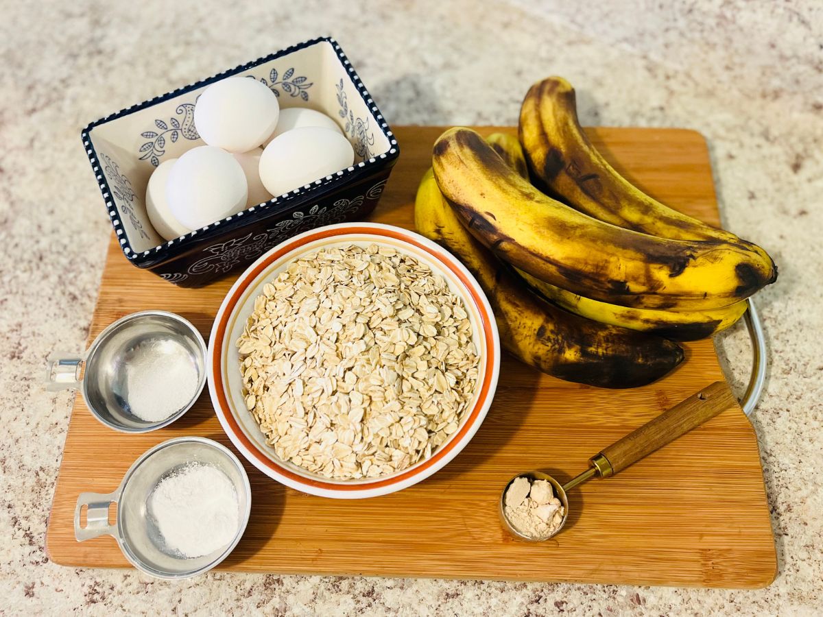 eggs in a ceramic container, ripe bananas, rolled oats in a bowl, salt in a silver plate and baking powder in a silver plate, sugar in a tablespoon, on a cutting wood board.