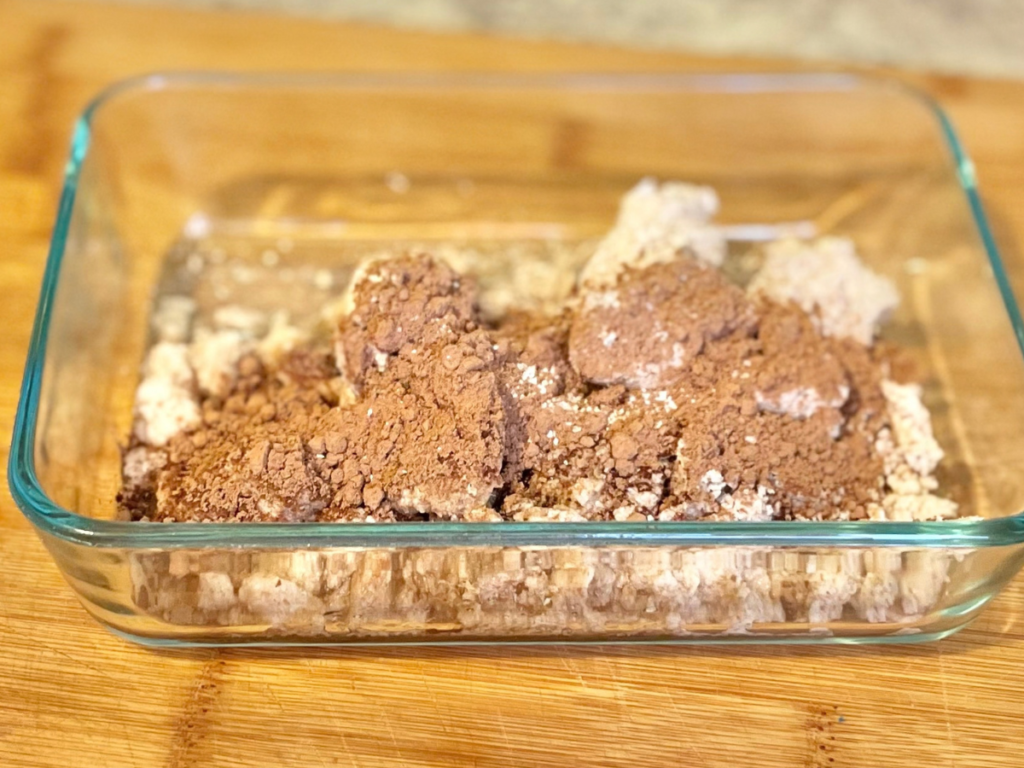 glass container with almond pulp and topped with cacao powder on top of a cutting board