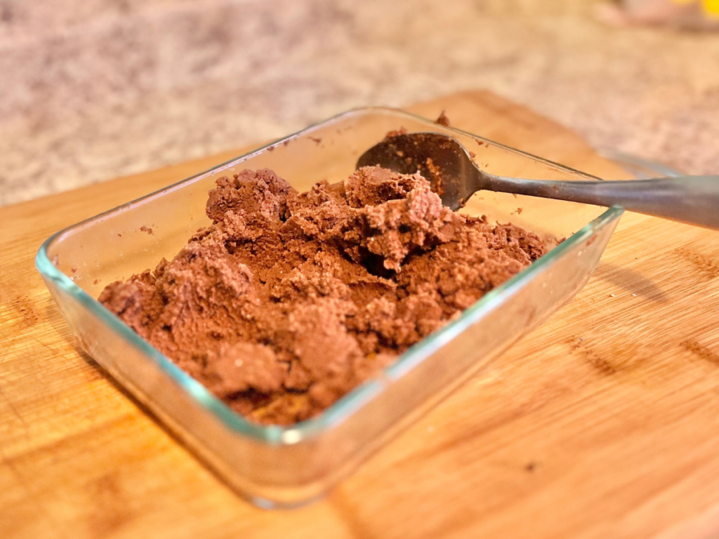 Glass container with brown chocolate spread with a silver spoon on a cutting board.