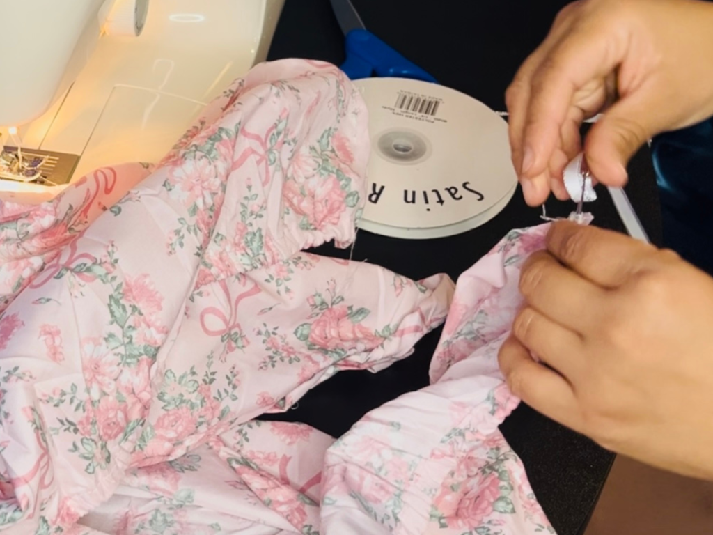 sewing machine next to pile of floral light pink fabric, threading with safety pin, and ribbon roll to the side with a hand holding pin.